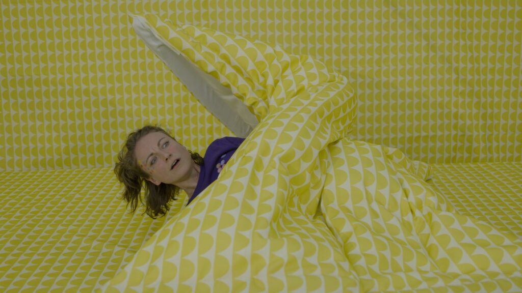 Eine Frau mit schulterlangen offenen Haaren schaut unter einer Decke hervor. Die Decke und der Hintergrund sind gelb gemustert.