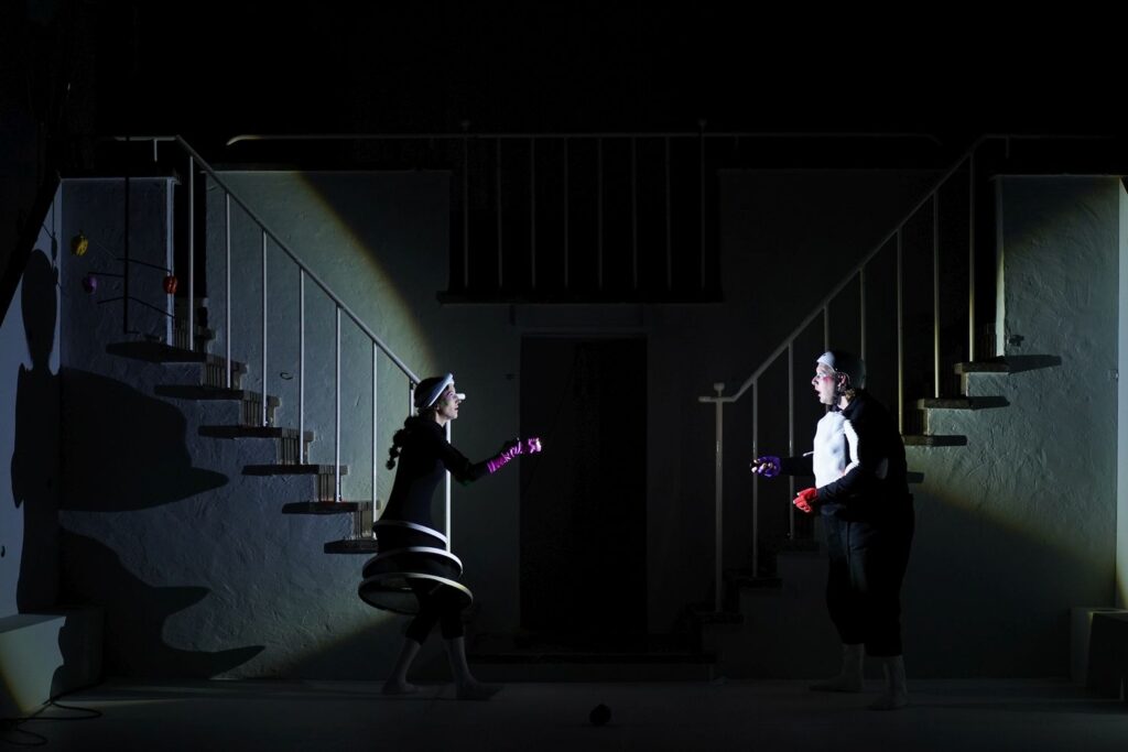 Cuyén Biraben und Lasse Wagner stehen sich in einem dunklen Raum gegenüber. Sie strahlen sich gegenseitig mit Taschenlampen an. Im Hintergrund sind zwei symmetrische Treppenaufgänge zu sehen.