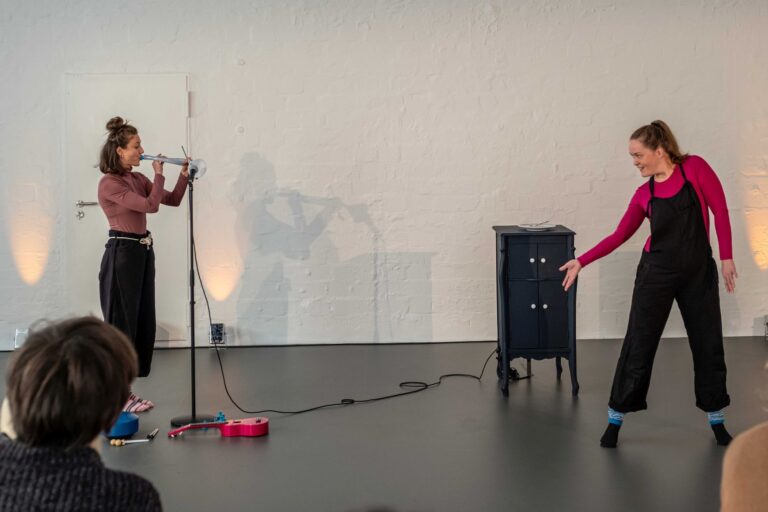 Links im Bild bläst eine Frau in einen Schlauch. Rechts im Bild macht eine Frau eine ausladende Bewegung mit der rechten Hand. Ihr Körper ist nach vorne gedreht. Bdeie Frauen tragen rosa Oberteile und schwarze Hosen.