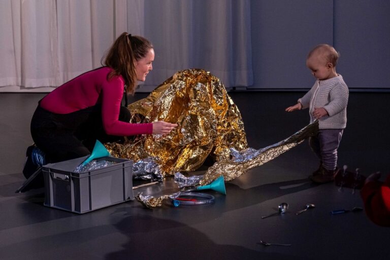 Links hockt eine Frau und blickt zu dem ca. 1-jährigem Kind rechts im Bild. Das Kind greift nach einer Rettungsdecke.