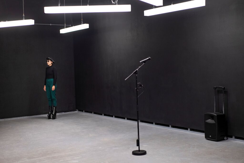 In einem leeren schwarzen Raum steht im Vordergrund ein Mikrofonständer. Im Hintergrund steht eine Frau in dunkler Kleidung und sehr hohen High-Heels.
