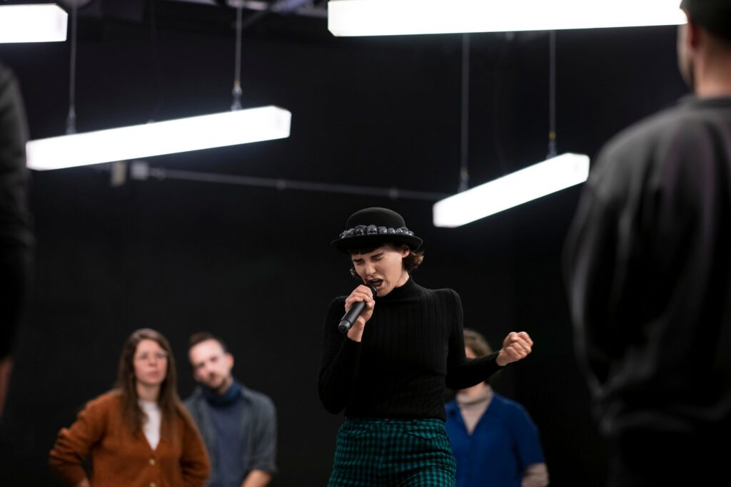 Lisa Bräuniger, eine schlanke weiße Frau mit kinnlangen dunklen Haaren, spricht mit großer Anspannung in ein Mikrofon. Über ihr hängen unsymmetrisch drei schmale, lange Deckenleuchten. Im Hintergrund stehen drei Menschen und schauen sie an.