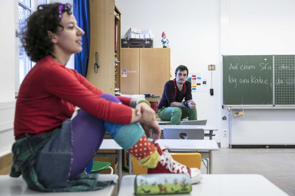 Eine Frau sitzt mit angezogenen Beinen auf einem Tisch in einem Klassenzimmer. Ein Mann sitzt neben der Tafel und beobachtet sie.