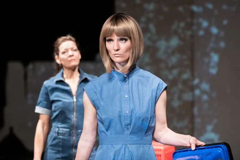 Eine Frau mit blonden glatten Haaren und blauem Kleid hält einen Einkaufskorb rechts neben ihrem Körper. Sie blickt durchdringend links an der Kamera vorbei. Hinter ihr steht eine Frau mit zusammengebundenen Haaren, die sie anschaut.