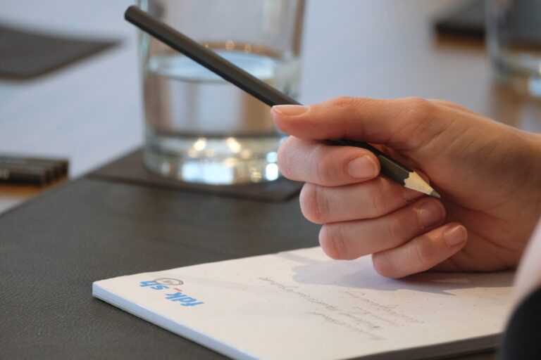 Eine Hand, die einen Bleistift hält, ruht auf einem beschriebenen Block. Oben rechts auf dem Block ist fdk_sh aufgedruckt.