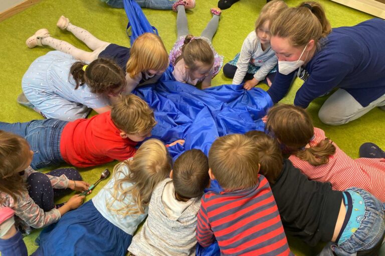 11 Kinder liegen bäuchlings im Kreis. In ihrer Mitte liegt ein blauer Stoff. Sie haben ihre Hände unter den Stoff gelegt. Anne sitzt mit den Kindern im Kreis und hält ihre Hände ebenfalls unter den Stoff. Sie trägt eine weiße FFP2-Maske.